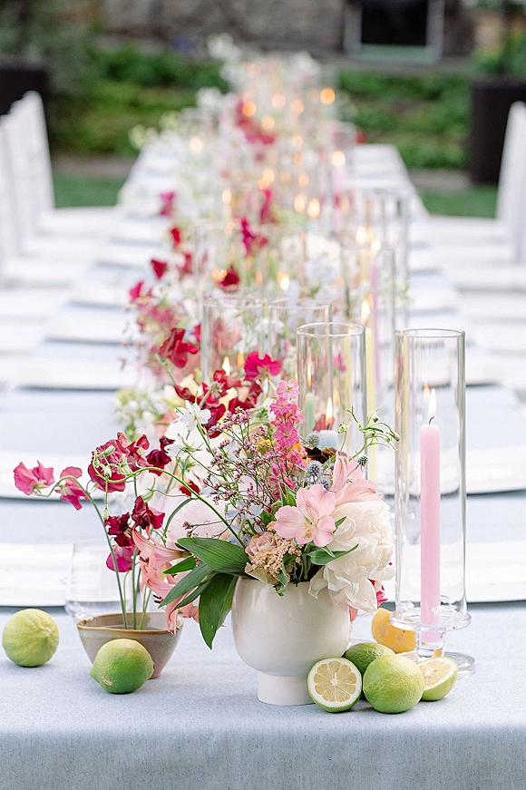 Reception tablescape with a long table centerpiece of wildflowers in ceramic vases, pastel taper candles, and citrus on light blue linens outdoors
