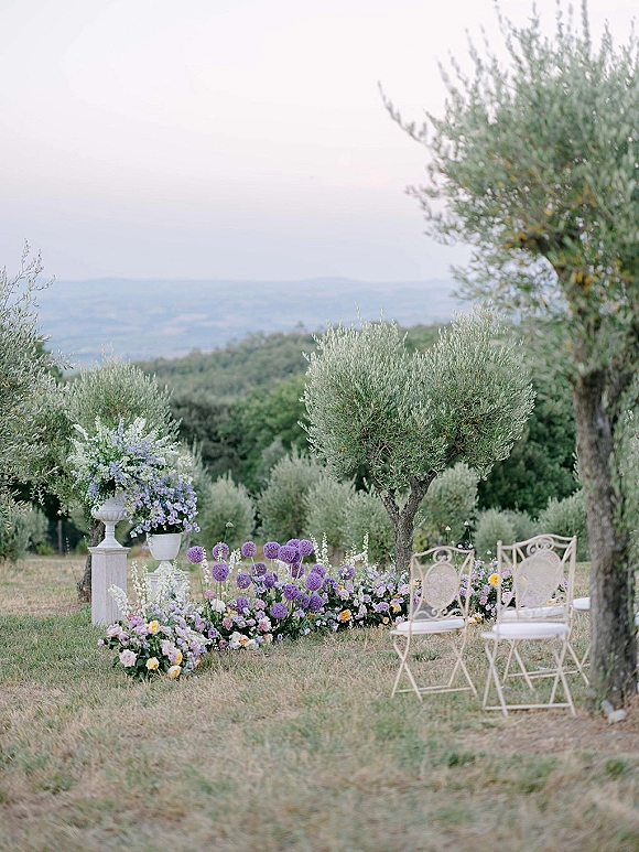 Ceremony setup with purple and white florals lining the aisle and white chairs, set in an olive grove with rolling hills beyond