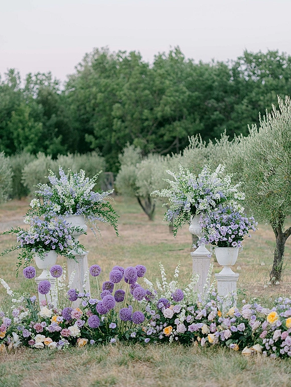 Wedding ceremony flowers in white urn planters on pedestal stands, with purple allium and pastel blooms lining an olive grove aisle entrance