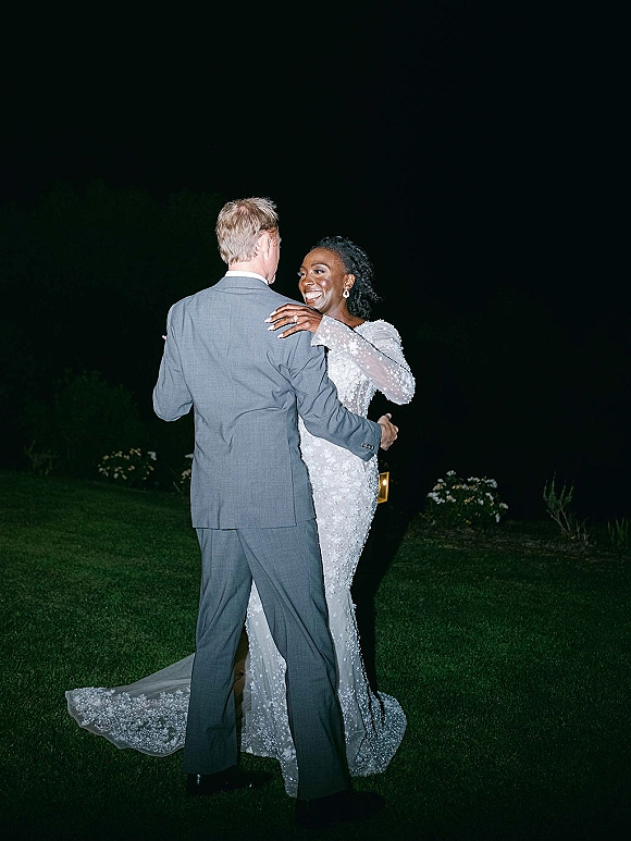 First dance outdoors as bride in a beaded gown with long train hugs groom in gray suit under night sky by lit garden path