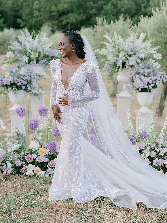 Bridal portrait of a smiling bride in a beaded wedding dress with cathedral veil and drop earrings in a lush garden setting