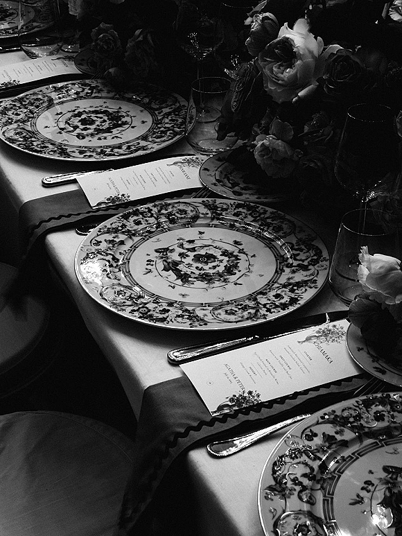 Reception tablescape with floral china wedding table settings, printed menu cards on plates, glassware, silver flatware, and a low rose centerpiece