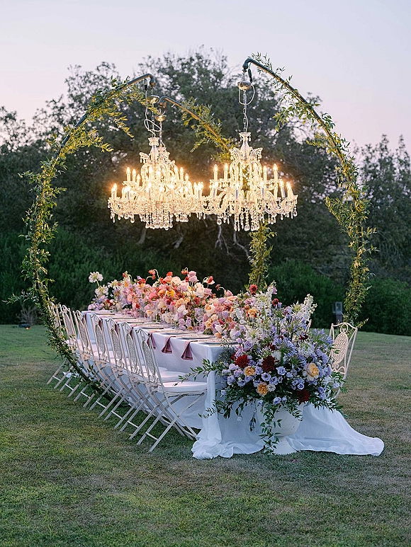 Reception tablescape with an outdoor reception table, long banquet linens, floral meadow runner, taper candles, and crystal chandeliers at dusk on a lawn