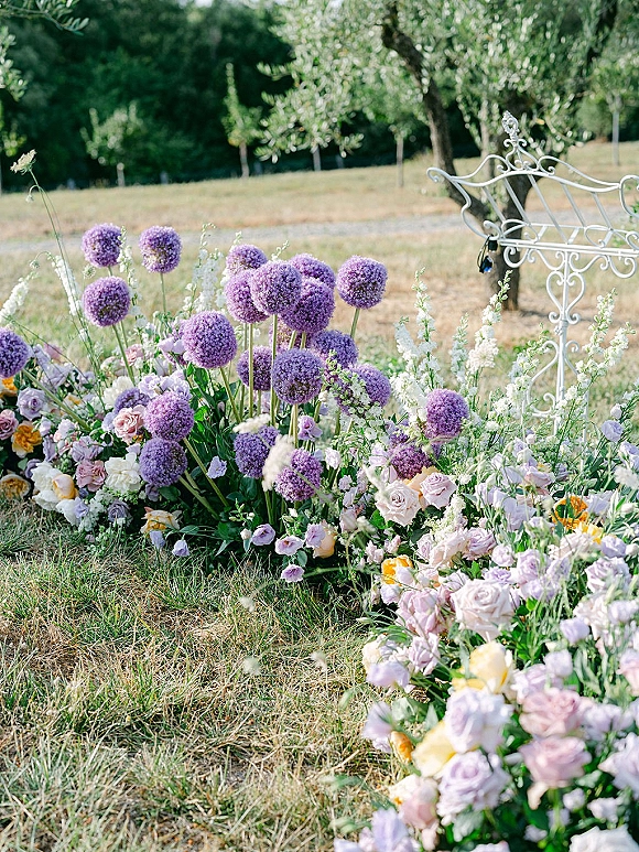 Ceremony aisle florals with purple allium and lavender roses in a meadow-like border along the grass lawn, with a white wrought iron chair nearby