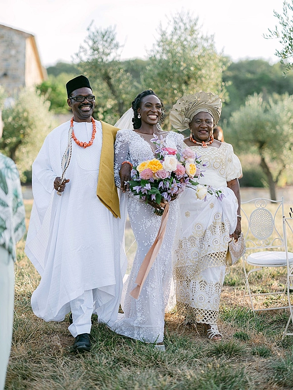 Bride walking aisle holding bouquet with trailing ribbon, veil and beaded long-sleeve gown in an outdoor garden beside a stone building