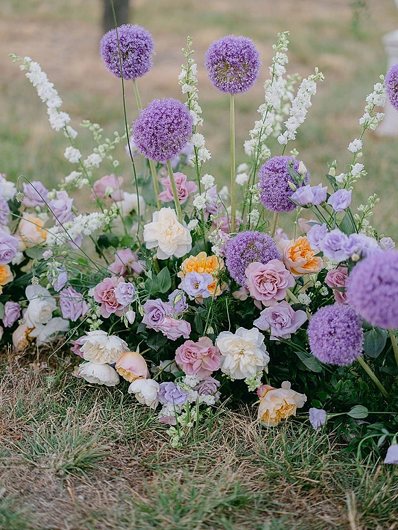 Wedding floral arrangement with purple allium and lavender and peach roses, nestled in greenery on a grassy outdoor field lawn