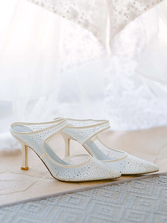 Bridal shoes with rhinestone accents, pointed-toe wedding heels resting by a bridal gown on an upholstered chair near a tiled floor