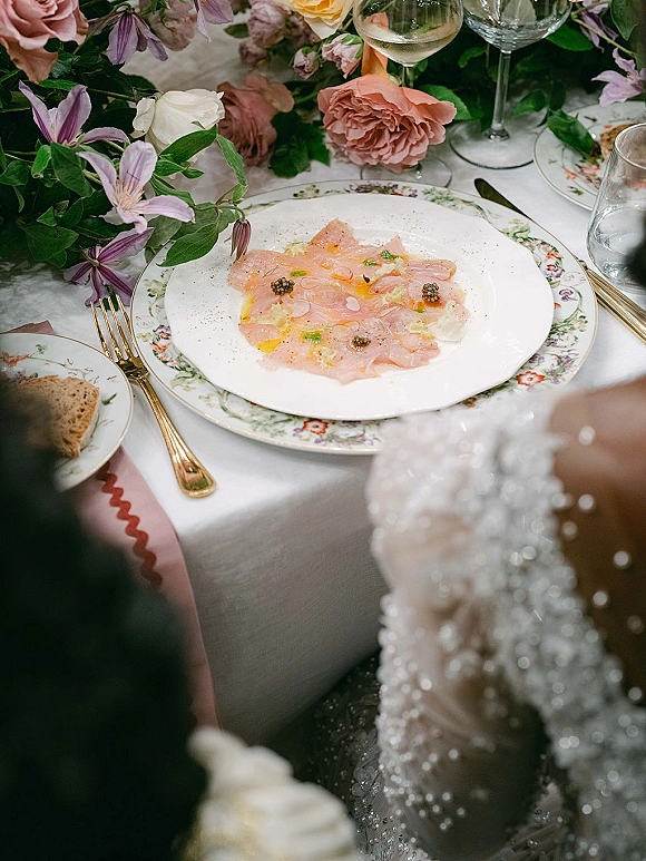 Wedding tablescape with vintage wedding table setting featuring rose and clematis centerpiece, patterned china, gold flatware, and wine glasses on linen cloth