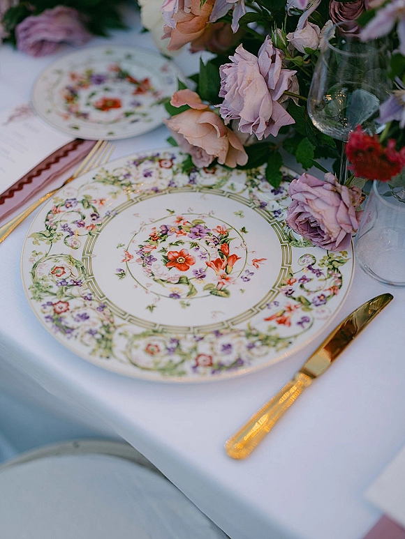Reception tablescape with vintage china wedding plates, gold flatware, and a pastel rose centerpiece on white linen in soft outdoor light