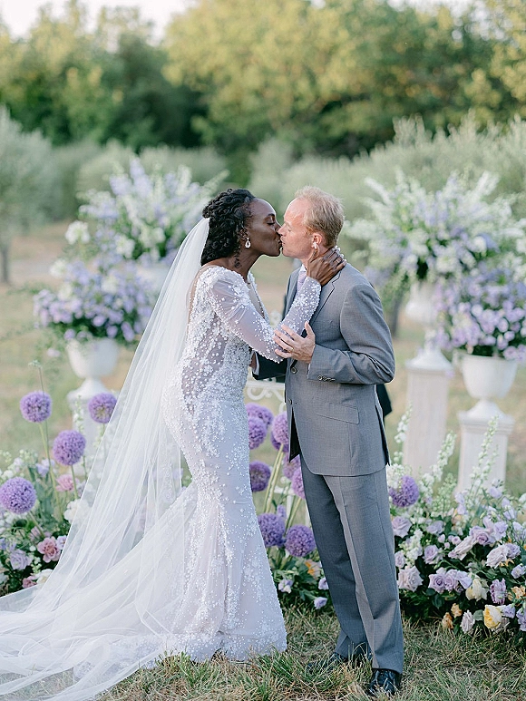 Wedding kiss portrait of bride and groom kissing, her long veil and beaded sleeves framed by lavender florals and garden greenery