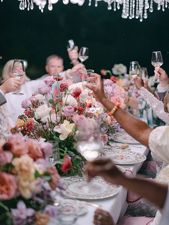 Wedding toast as guests raise wine glasses over a candlelit dinner table with floral centerpiece beneath a crystal chandelier in an outdoor tent at night