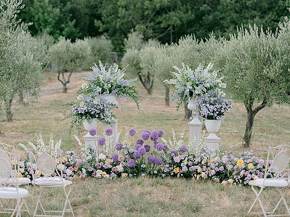 Ceremony altar decor with outdoor wedding altar flowers in white urns and pedestal stands, lavender-purple blooms, greenery, and olive trees behind