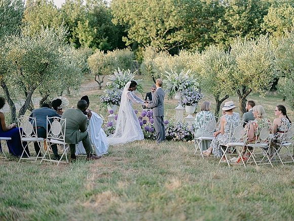 Wedding ceremony with bride and groom under a flowered arch, long veil trailing, guests seated among olive trees on a lawn