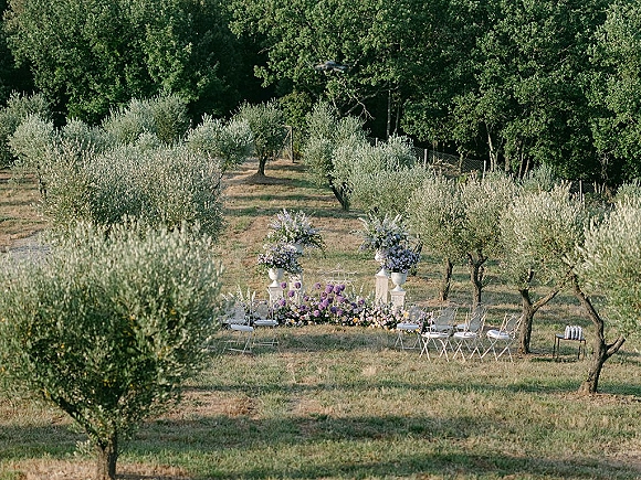 Outdoor ceremony setup with garden ceremony seating, white pedestals and floral urns framing an aisle runner in an olive grove field