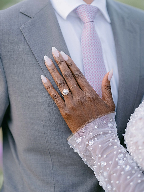 Engagement ring close-up of an oval diamond engagement ring on a nude-manicured hand resting on groom’s suit chest with blurred greenery behind