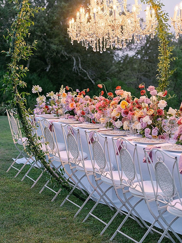 Reception tablescape with a long banquet table, pastel rose centerpieces and greenery garland, taper candles, under a crystal chandelier on a garden lawn