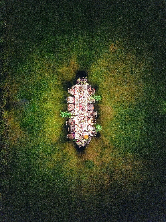Reception tablescape with an outdoor wedding dinner table featuring a long banquet setup, floral centerpieces, and taper candles on a lawn