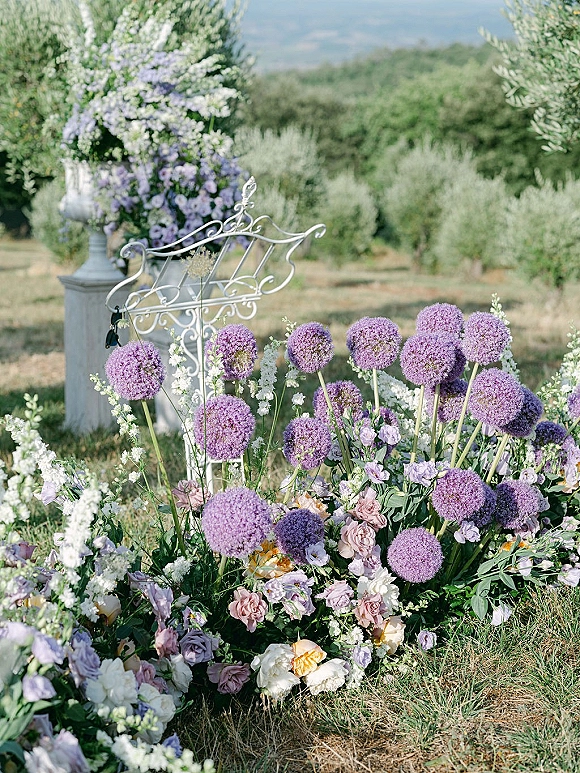 Wedding ceremony flowers with a ceremony floral ground arrangement of pastel roses and purple allium beside a white music stand on a grassy lawn