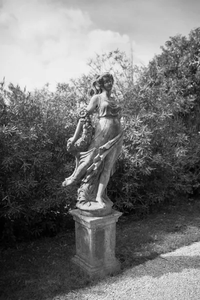 Garden statue stone garden sculpture on a pedestal base with floral garland detail beside a gravel path, shrubs, and trees under sky