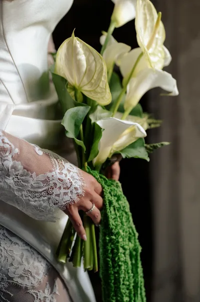 Bridal bouquet of calla lilies and white anthurium with cascading greenery held by a bride in a lace sleeve against a neutral wall