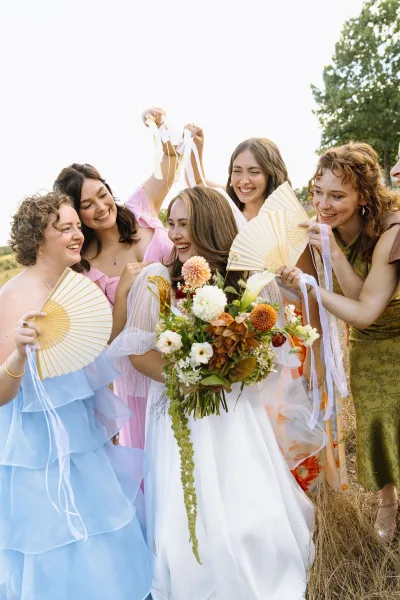 Bridal party portrait of a bride with bridesmaids holding ribbon wands in a grassy field, trees and sky behind.