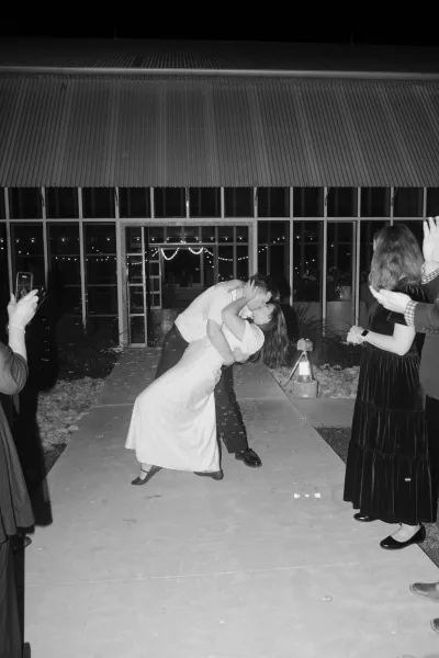 Wedding exit bubble send off as the couple shares a dip kiss under string lights along a walkway beside a glass facade at night.