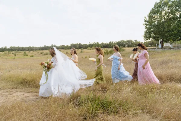 Bride and bridesmaids, a bridal party walking through meadow grass, laugh together beneath open sky, some carrying hand fans.