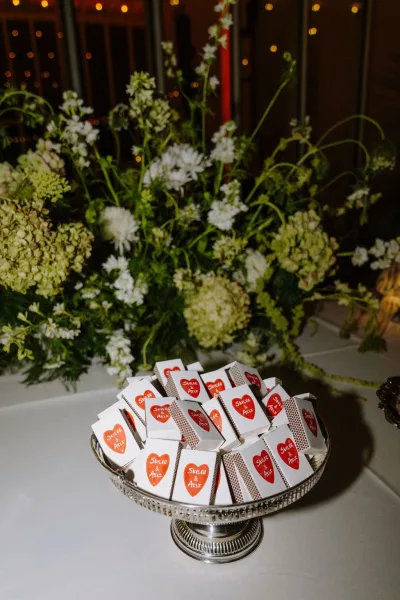 Matchbox wedding favors, personalized matchbooks neatly arranged on a silver tray, set before greenery and white flowers under string lights.