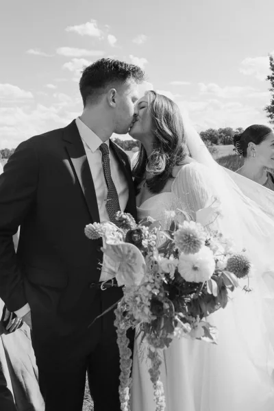 Wedding kiss portrait as a bride and groom kiss on a lakeside lawn, her veil drifting, trees and sky reflected on the calm water.