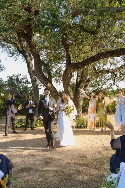 Ceremony recessional as the bride and groom exit under oak trees along a gravel path, wedding party cheering with handheld fans.