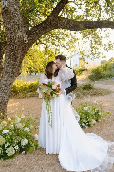 Wedding kiss portrait as the bride and groom embrace beneath an oak tree, the bride holding a rust and white bridal bouquet in golden light.