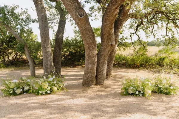 Ceremony altar decor with ground arrangements framing a gravel aisle in an outdoor ceremony setup under trees beside a meadow.