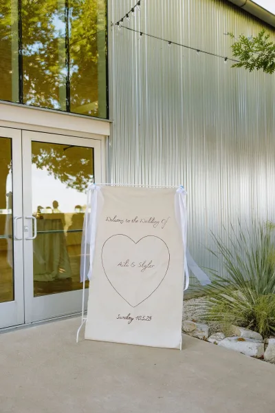 Wedding welcome sign on a fabric welcome banner with a heart motif, set before a corrugated metal wall and glass doors.