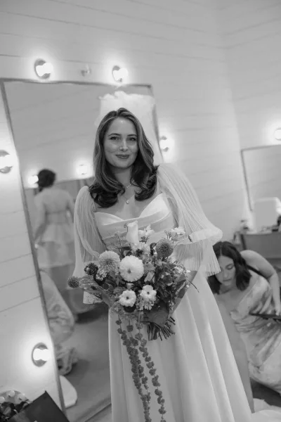 Bridal portrait of a bride getting ready, holding a bouquet in a dressing room with mirror and glowing vanity lights against shiplap.