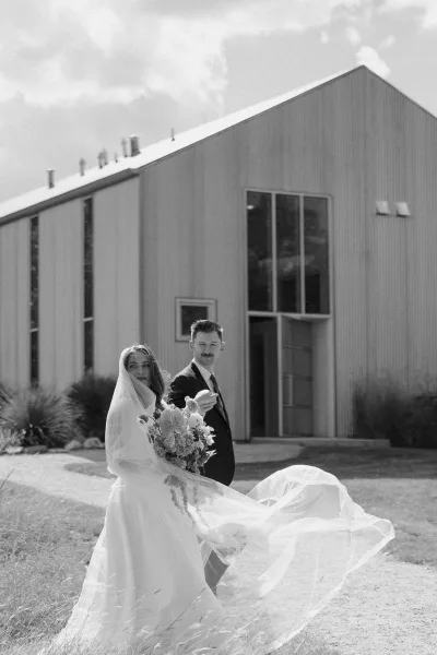Bride and groom walking in a black and white wedding photo along a gravel path by a rustic barn, her veil trailing at her side.