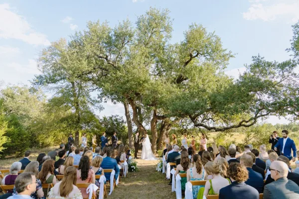 Ceremony moment under a large tree at an outdoor wedding ceremony, with wooden chairs lining the aisle on a sunlit grass lawn.