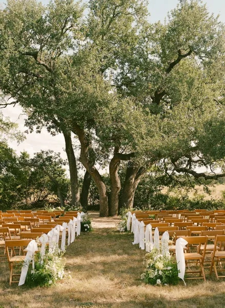 Outdoor ceremony setup for a ceremony under a large tree, with rows of wooden chairs framing a simple aisle on a grass lawn in an open field.