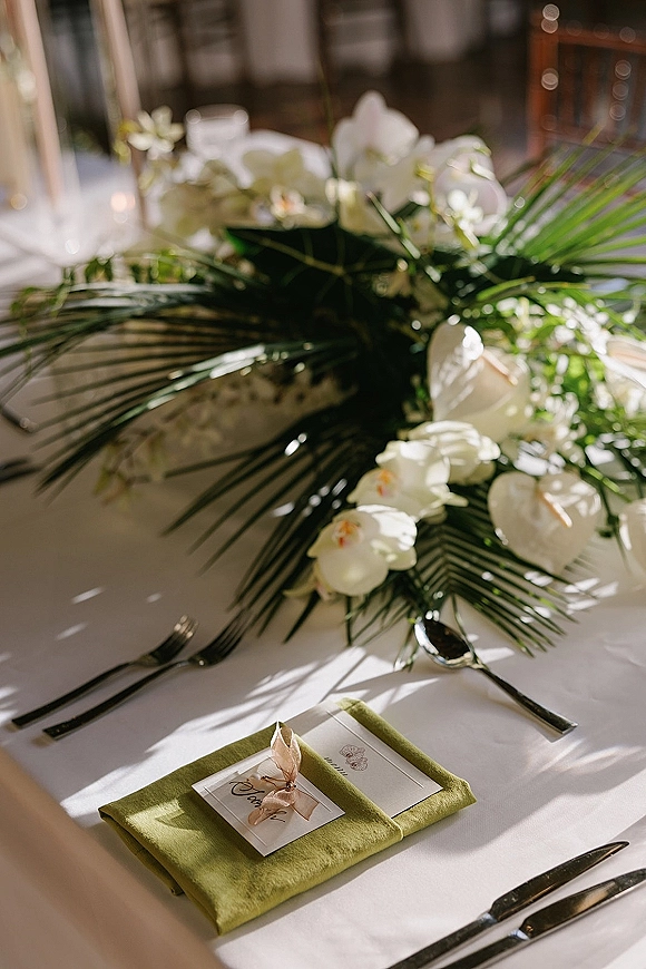 Reception tablescape with wedding place setting, green napkins and ribboned place cards, white orchids and palm fronds by window light with candles