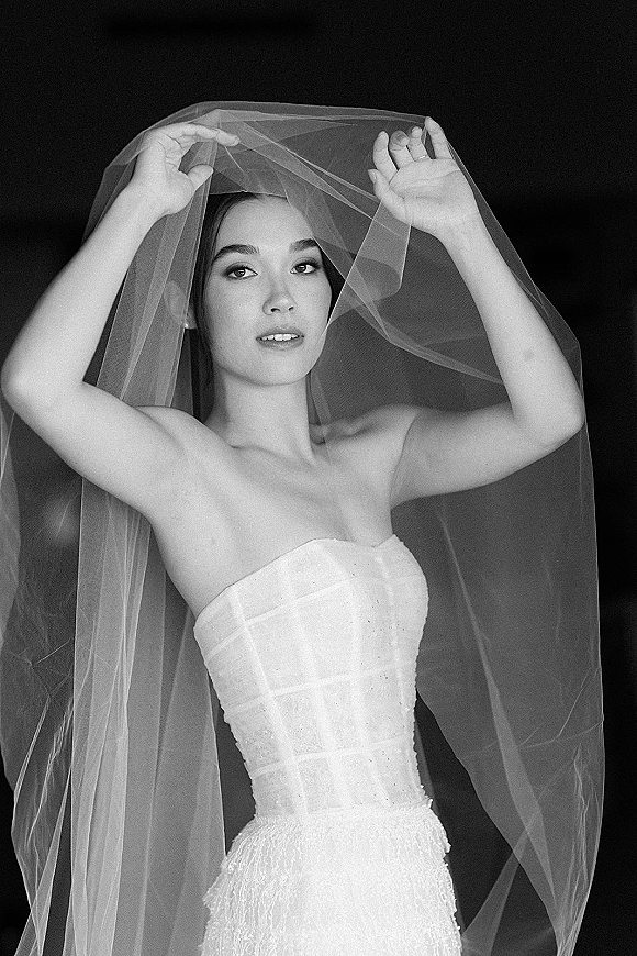 Bridal portrait in black and white of a bride holding her long sheer veil, wearing a strapless beaded gown and drop earrings against a dark studio backdrop