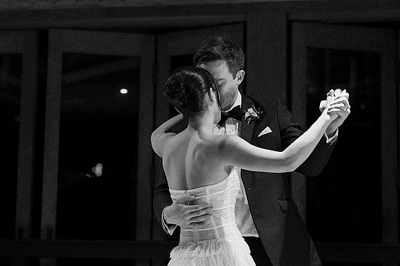 First dance as bride in a strapless wedding dress and groom in tuxedo embrace on an indoor reception floor, dark paneling behind