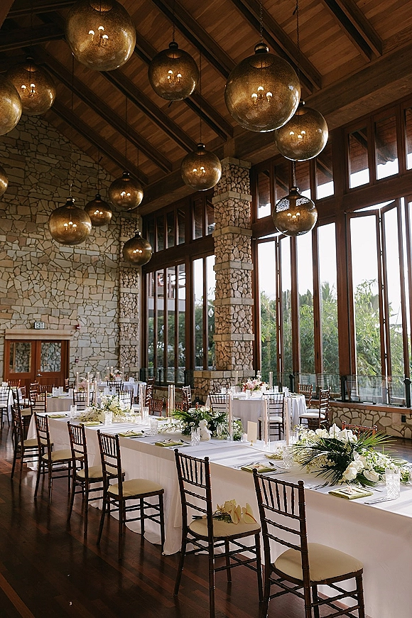 Reception tablescape with long banquet table wedding styling, greenery garlands, white florals, taper candles, and globe chandeliers in a stone-walled venue