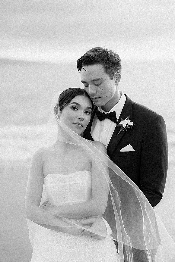 Couple portrait in a black and white wedding portrait, bride leaning on groom as he hugs her by the ocean horizon, veil flowing