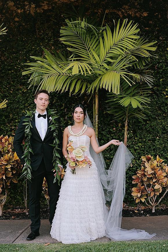 Couple portrait of bride and groom portrait in a tropical garden, bride in strapless gown and long veil holding bouquet beside tuxedo groom with leaf lei.