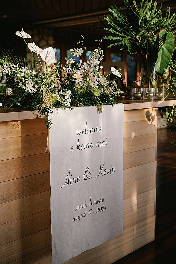 Wedding welcome sign with fabric banner and calligraphy lettering, framed by tropical greenery and white orchids on a wood bar in warm sunlight