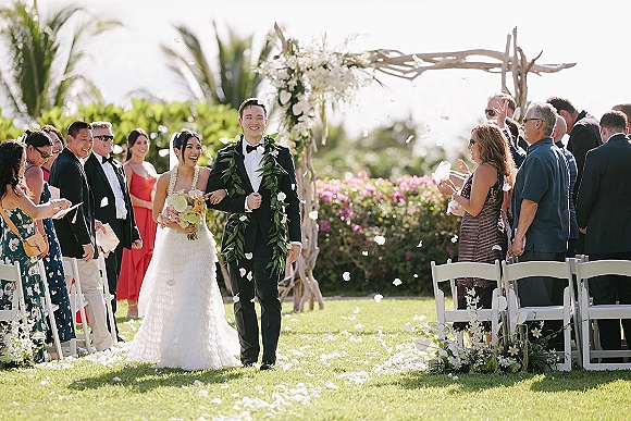 Wedding recessional as bride and groom walk aisle smiling, bouquet and black tuxedo amid flower petals, guests cheering under palm trees