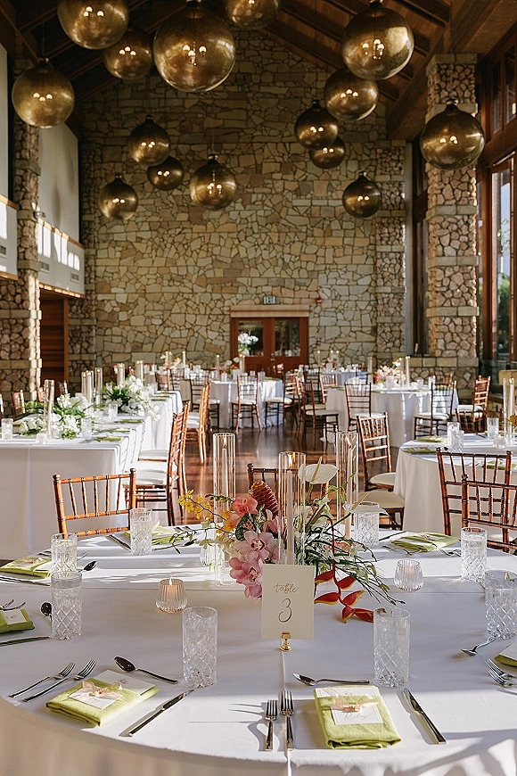 Reception tablescape with wedding reception table decor, white tablecloths, floral centerpieces, tall cylinder candles, and green napkins in a stone-walled hall