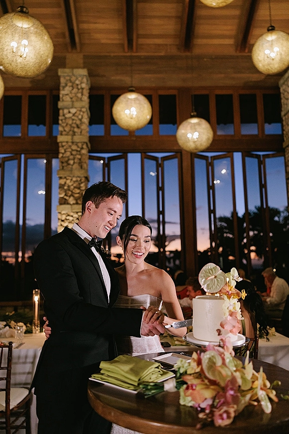 Wedding cake cutting as bride in a strapless gown and groom in a tuxedo slice a one-tier cake with tropical flowers at sunset-lit reception windows