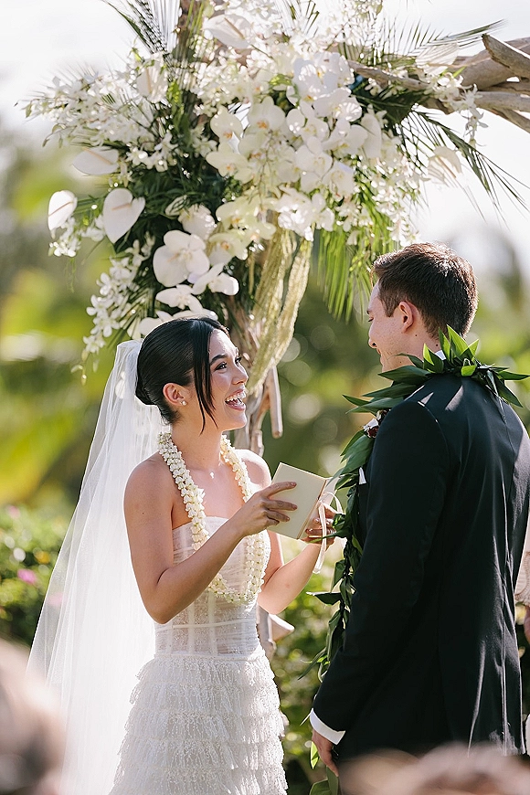 Wedding vows as the bride reads vows from a book to the groom under a white orchid arch in a sunlit outdoor garden ceremony