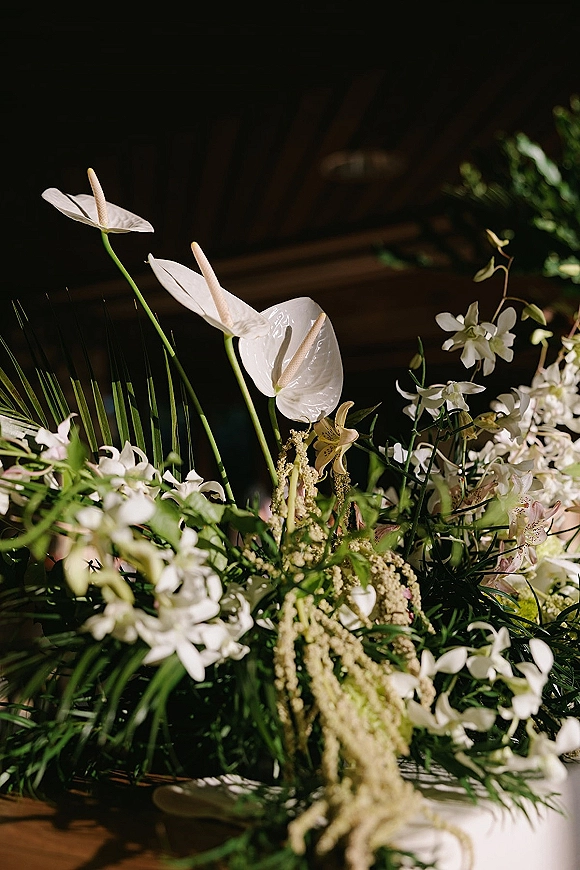 Wedding floral centerpiece with white anthurium and orchids, lush greenery and palm fronds arranged low on a wooden table indoors