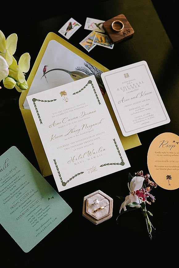 Wedding invitation suite flat lay with envelopes, RSVP and details cards, wax seal, vintage stamps, rings, and orchid on dark tabletop